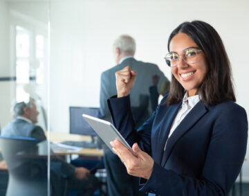 Happy female professional in glasses and suit holding tablet and making winner gesture while two businessmen working behind glass wall. Copy space. Communication concept